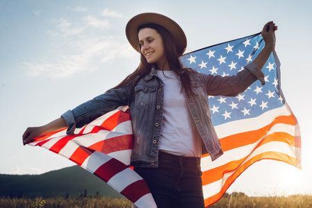 Portrait of beautiful young caucasian woman in hat standing on background blue sky and mountains in wind independent and young, holds proudly usa flag high in air, demonstrates patriotism and freedomの写真素材