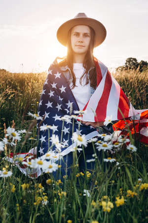 Calm young caucasian woman 20s in hat sitting on green grass wrapped in usa flag looking at camera outdoor. Independent confident brunette lady vertical headshot portrait. 4th of July conceptの写真素材