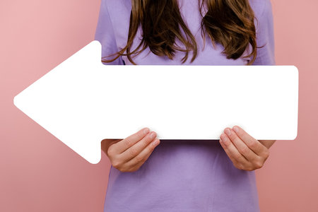 Close up cropped young woman holding big white paper arrow, posing isolated over pink studio background with copy space for promotional content. Business vision, achievement and target conceptの写真素材