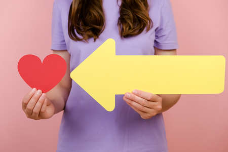 Close up of unrecognizable young female holds little red hearts and yellow arrow, isolated on pink...
