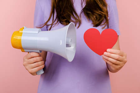Close up cropped young woman holding small paper red heart and purple megaphone, dressed t-shirt, posing isolated over pink color background in studio. Valentine's day and tenderness conceptの写真素材