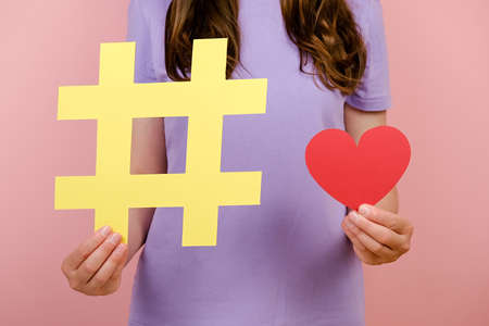 Close up cropped footage of young woman wears t-shirt demonstrates large big yellow hashtag sign and little red heart, posing isolated over pink background in studio, symbol of charity on Internetの写真素材