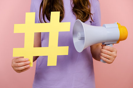 Close up cropped young woman holding yellow hashtag sign and megaphone, label for business, marketing and advertising, isolated on pink background. Concept social network monitoring, media measurementの写真素材