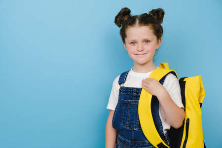 Portrait of cute beautiful happy smiling caucasian little girl, kid schoolgirl holding yellow backpack standing over blue studio background wall, positive looking at camera. Back to school conceptの写真素材