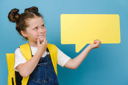 Portrait of thoughtful smiling little caucasian schoolgirl holding yellow blank speech bubble above...