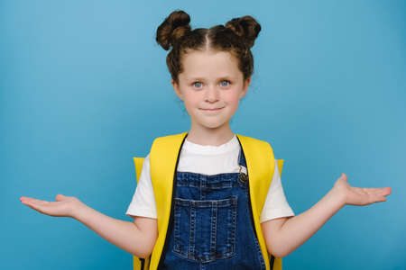 Portrait of lovely sweet small schoolgirl wears yellow backpack raise two palms arms holding empty space beaming shiny smiling positive good look, posing isolated over blue color background in studioの写真素材