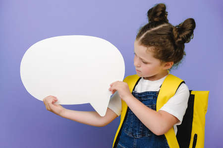 Cute elementary schoolgirl holding white speech bubble, wears yellow backpack, isolated over purple studio background with copy space for promotion consent. Thinking of school. mockup, free spaceの写真素材
