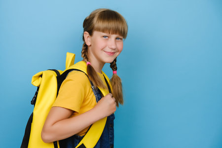 Happy smiling schoolgirl with yellow backpack bag standing isolated on blue studio background with copy space for advertisement, positive looking at camera. Portrait of smart caucasian kid studentの写真素材