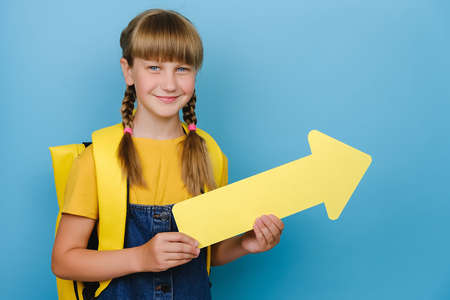 Portrait of smiling blonde schoolgirl showing aside with yellow arrow, wears backpack, posing isolated over blue color background wall with copy space for promotional content. Back to school conceptの写真素材