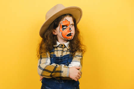Close up portrait of angry sad little girl child with halloween makeup mask, wears brown hat and...
