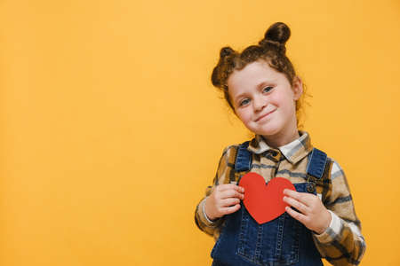 Portrait of positive little sincere adorable child girl holding red heart on chest feeling gratitude, isolated over yellow studio background, gesture of love appreciation gratitude. Health day conceptの写真素材