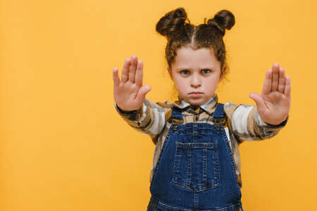 Portrait displeased little kid girl demonstrate stop gesture, wears shirt, posing isolated over yellow color background studio with copy space for advertisement. Childhood emotional lifestyle conceptの写真素材