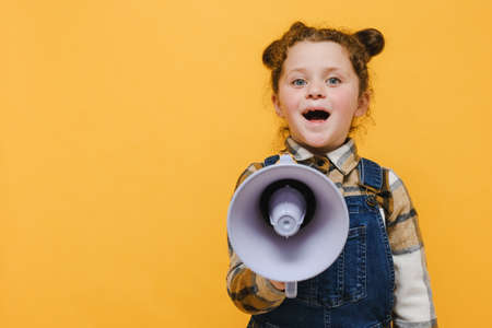Cheerful cute funny little child girl screaming in megaphone, posing isolated over pastel yellow color background studio with copy space for promotion content. Childhood emotion lifestyle conceptの写真素材