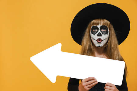 Scary bright alive girl child with Halloween makeup mask showing aside with white arrow wears big...