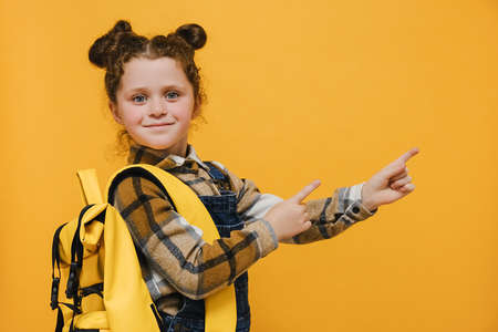 Closeup portrait of nice cute attractive happy glad schoolgirl wear yellow backpack pointing two fingers looking aside copy space for advertisement, posing isolated over yellow color background studioの写真素材