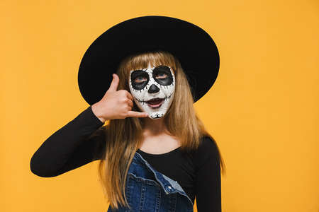 Portrait of girl child with Halloween makeup mask in black hat do phone gesture like says call me back, posing isolated over yellow color background wall in studio. Celebration holiday party conceptの写真素材