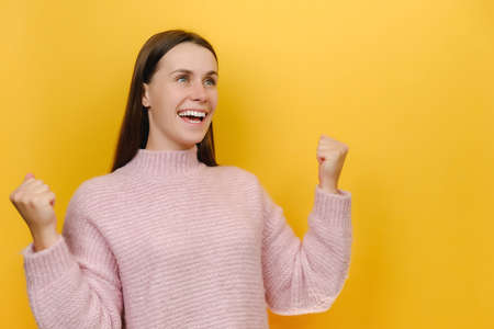 Excited happy successful overjoyed lucky attractive young caucasian woman 20s doing winner gesture celebrating clenching fist say yes, wears pink sweater, isolated on yellow color background studioの写真素材