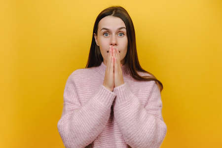 Shocked young caucasian woman 20s hold arms on mouth hiding amazed facial expression bad news epic fail made mistake wear casual pink sweater, posing isolated yellow color background in studioの写真素材