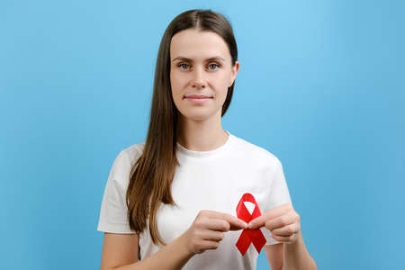 Young brunette woman 20s holds red ribbon, looking at camera, isolated on blue color background studio. Blood transfusion and donation. Hemophilia and World Health concept. World AIDS and hiv dayの写真素材