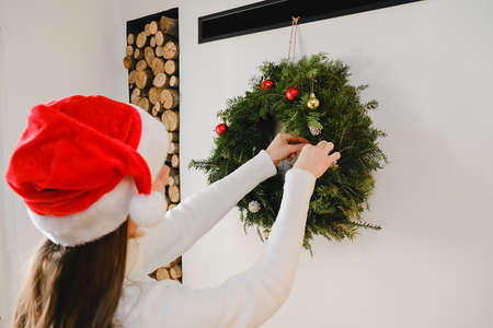 Back view of young caucasian woman 20s old years in Santa Claus hat decorated Christmas wreath with New Year decorations, standing near fireplace in living room at home. Xmas winter holiday conceptの写真素材