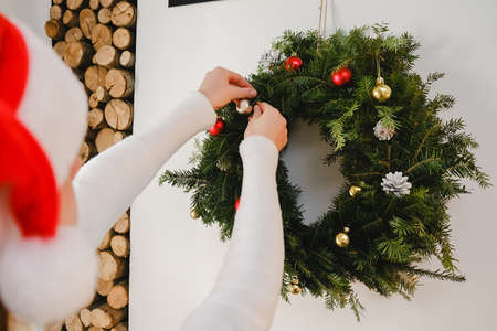 Selective focus of young woman wearing sweater and santa red hat decorating Christmas wreath with toys and balls at home, preparing home for New Year celebration, enjoying holiday, festive interiorの写真素材