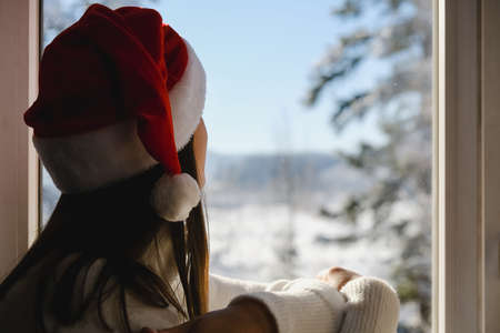 Selective focus of young woman in stylish white knitted sweater and Santa red hat sitting at home on windowsill looks at the beautiful snowy mixed forest. Dreamy cute girl enjoys holiday weekendの写真素材
