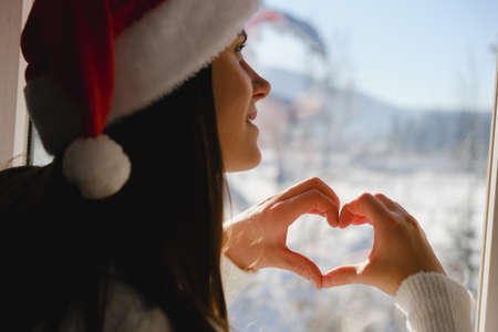 Back view of smiling young Santa girl in white sweater and Christmas hat Showing shape heart with...