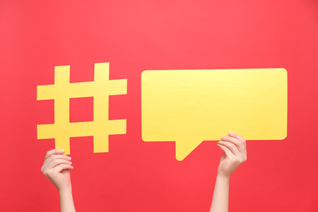 Female hands holding yellow paper hashtag symbol and speech bubble, hash sign of famous media content, isolated on red color background. Post viral web network, blogging blog website strategy conceptの写真素材