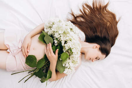 Future mother's expectation. Head shot portrait young pregnant woman with perfect skin touching gently tummy hand lying on comfortable bed with beautiful fresh summer flowers. concept motherhoodの写真素材