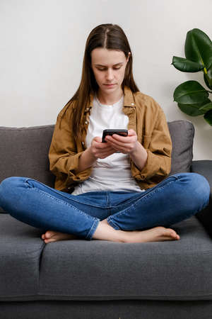 Beautiful calm young woman using smart phone sitting on comfortable sofa at home. Attractive girl...