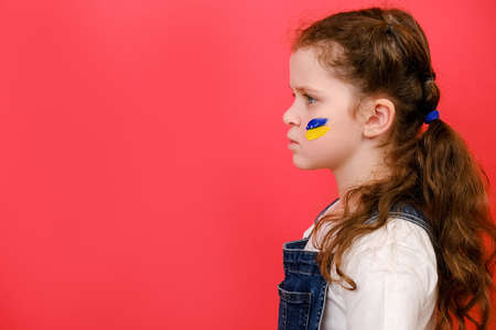 Profile side view of little girl with flag of ukraine on face, posing isolated on red studio background with copy space for advertisement. Peace, no war, help, stop russian aggression. crisis conceptの写真素材