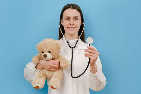 Portrait of cute smiling young woman doctor holding stethoscope and small fluffy bear, looking at camera, isolated over blue color background wall in studio. Pediatrician with toy. medical conceptの写真素材