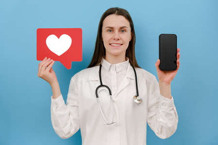 Cute young woman physician practitioner in white uniform with stethoscope holds heart icon and...