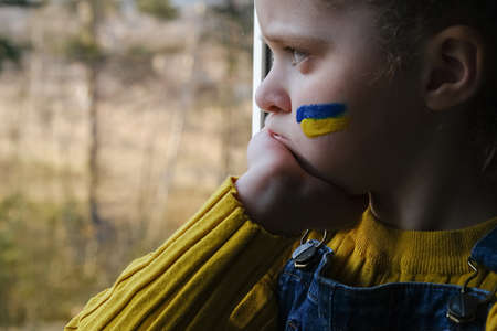 Close up of sad small girl kid with flag of Ukraine on face sitting alone in windowsill at home. Unhappy child on nature background. Peace, no war, help, stop russian aggression. crisis conceptの素材 [FY310185381552]