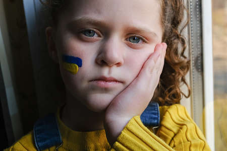 Sad little Ukrainian girl with flag of Ukraine on her face sitting on windowsill at home. Children are asking for peace. Concept of participation of Ukrainian people in war with Russia. no warの写真素材
