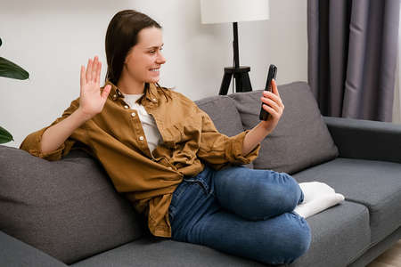 Smiling young female sitting on couch waving hand using smartphone app enjoying online virtual chat...