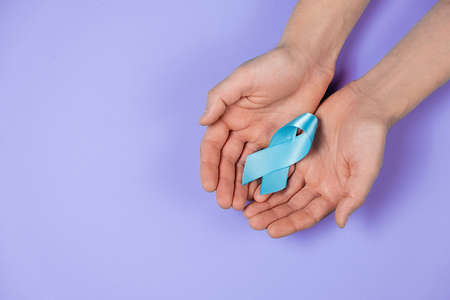 Close up top view of male hands holding small blue awareness ribbon on purple color background wall in studio with copy space for advertisement. Symbol of social and medical issuesの写真素材