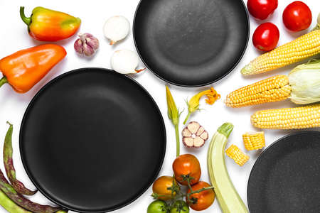 Creative layout made of autumn vegetables. Tomatoes, onion, carrot, garlic, red beet, pepper, zucchini, corn, green haricot and black plate on white studio background. Flat lay, top view, copy spaceの写真素材