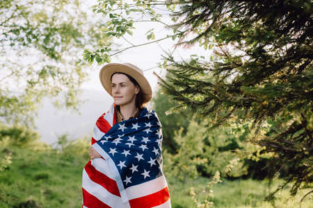 Portrait of patriotic young female in beige hat wrapped big usa United States flag standing on background mountains during sunset. Hipster girl feeling freedom, independence concept. July 4th conceptの写真素材