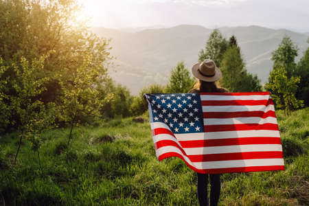 Back view of patriot young woman in hat standing on background summer mountains during beautiful amazing sunset or sunrise, holds proudly usa flag high in air, demonstrates patriotism and freedomの写真素材