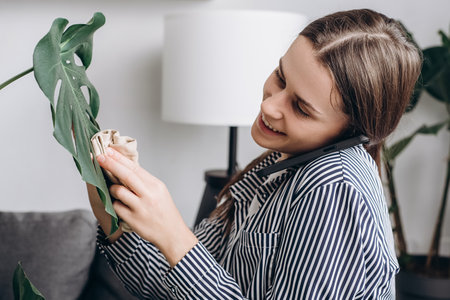 Smiling young woman gardener caring houseplants in greenhouse garden. Happy female plant shop owner taking order from customer on mobile phone. Small business entrepreneur and plant caring conceptの写真素材