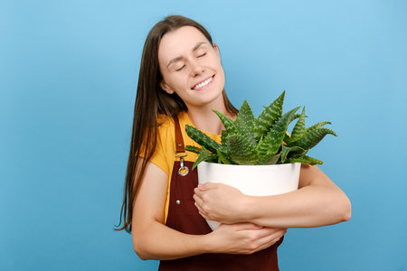 Dreamful young female closes eyes embraces pot of houseplant, new flower for home garden, posing isolated over blue color background wall in studio. Gardening, housewife, people and lifestyle conceptの写真素材