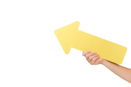 Close up of female hand holding big yellow paper arrow pointer on copy space for promotion content or design, posing isolated over white studio background wall. business and strategic conceptの写真素材