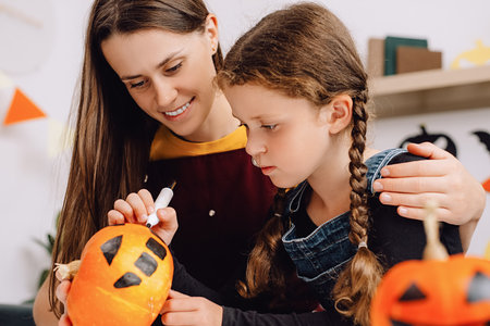Selective focus of loving young mother teaches cute daughter painting on orange pumpkin sit together on couch at home, smiling parent and little kid preparing handmade decorations. halloween conceptの写真素材