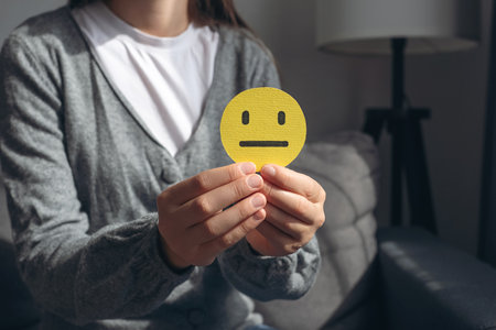 Unrecognizable young woman holding small yellow sad face, demonstrate bad emoticon sitting on gray couch. Feedback rating and positive customer review, experience, satisfaction survey conceptの写真素材
