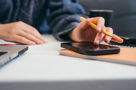 Close up of female hands making notes on smartphone and in notebook, planning work calls. Student...