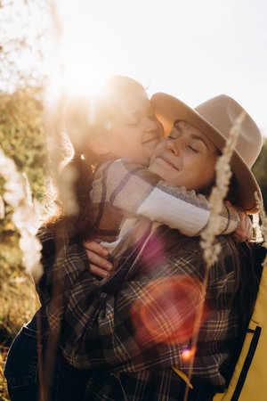 Vertical shot of loving pretty little daughter hugging with smiling young mother sit in grass on background beautiful autumn forest during epic sunset or sunrise. Concept family relations and travelの写真素材