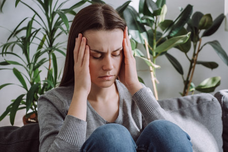 Upset depressed young girl sitting on sofa feeling strong headache migraine, sad tired drowsy...