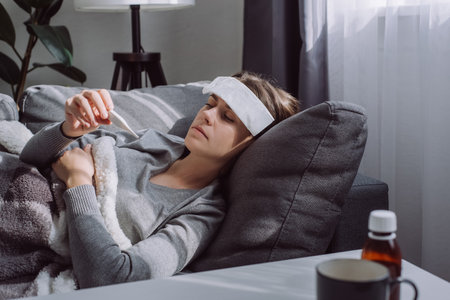 Woman under plaid feel sick unwell. Unhealthy young female measuring body temperature with electronic thermometer, suffer from flu lying on sofa at home, seasonal contagious grippe illness conceptの写真素材