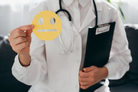 Close up of woman doctor in white uniform with stethoscope holding paper yellow sad face, bad smiley sitting on sofa. Feedback rating and customer review, experience, satisfaction survey conceptの写真素材